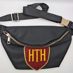 May include: Black fanny pack with a gold zipper and buckle. The pack features a maroon and gold patch with the letters "HTH". The adjustable strap is black.