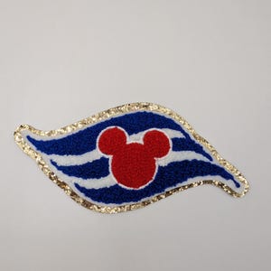 May include: A fabric patch in a stylized flag shape, with blue and white stripes, a red Mickey Mouse head, and a gold glitter border. Suitable for clothing or accessories.