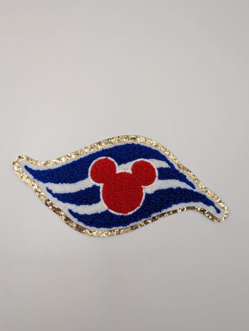 May include: A fabric patch in a stylized flag shape, with blue and white stripes, a red Mickey Mouse head, and a gold glitter border. Suitable for clothing or accessories.