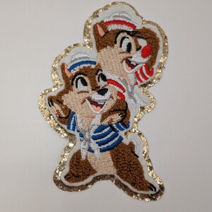 May include: Embroidered patch depicting two cartoon chipmunks, one in a blue and white striped sailor shirt and the other in a red and white striped shirt. The patch has a gold glitter border on a white background.