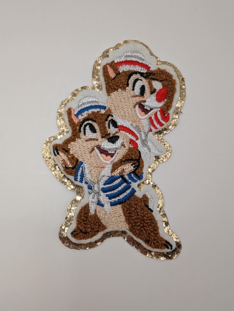 May include: Embroidered patch depicting two cartoon chipmunks, one in a blue and white striped sailor shirt and the other in a red and white striped shirt. The patch has a gold glitter border on a white background.