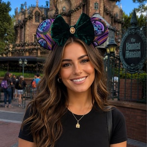 Op de afbeelding: Een persoon draagt een zwarte top en een Haunted Mansion-thema oorband met een groene fluwelen strik. De achtergrond toont de gevel van Haunted Mansion en een bord met de tekst "The Haunted Mansion".