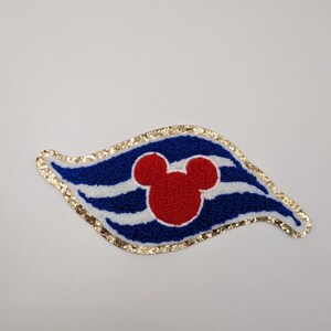 May include: A decorative patch with a red Mickey Mouse head on a blue and white striped design. The patch has a gold glitter border and is shaped like a stylized flag.