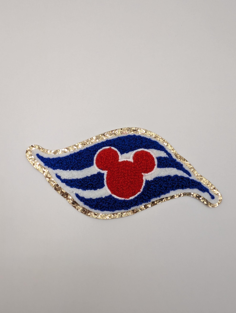 May include: A decorative patch with a red Mickey Mouse head on a blue and white striped design. The patch has a gold glitter border and is shaped like a stylized flag.
