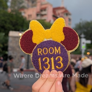 Może przedstawiać: Naszywka w kształcie Myszki Minnie z żółtą kokardą, bordowymi uszami i fioletowym środkiem. Na środku napis "ROOM 1313" złotą nicią. Naszywka ma złotą, błyszczącą obwódkę i tekst "Where Dreams Are Made" poniżej.