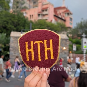 May include: A maroon shield-shaped patch with gold embroidered text "HTH" and a gold border. The patch is held up in front of a pink building. The text "Where Dreams Are Made" is written on the image.