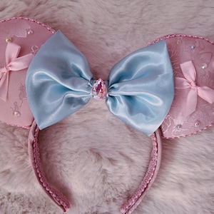 May include: Pink mouse ear headband with a light blue satin bow and a pink jewel. The ears are embellished with small pink bows and rhinestones. The headband is pink and has a textured design.