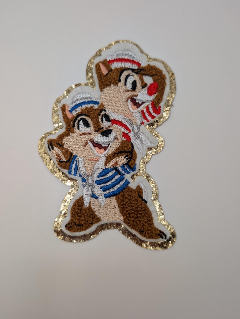 May include: Embroidered patch featuring Chip and Dale, Disney characters. The patch shows the chipmunks in sailor outfits, with brown fur, white hats, and blue and white striped shirts. The patch is outlined with gold sequins.