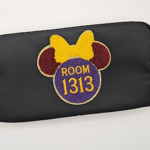 Może przedstawiać: Czarna prostokątna kosmetyczka z naszywką z motywem Myszki Minnie. Naszywka ma żółtą kokardę, bordowe uszy i fioletowe koło z napisem "ROOM 1313" w kolorze złotym. Torba jest wykonana z gładkiego, matowego materiału.