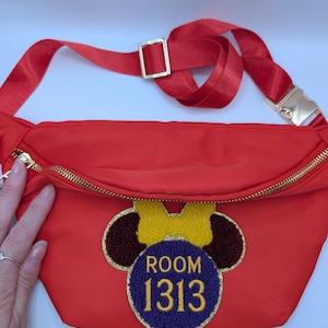 May include: Red fanny pack with a gold zipper and adjustable strap. The pack features a Minnie Mouse-inspired design with the text "ROOM 1313" in a circular patch. The strap has a gold buckle.