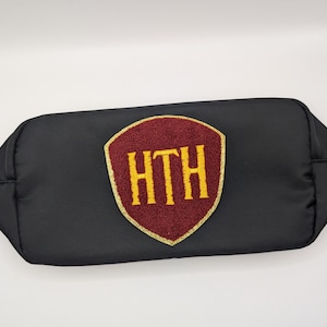 Może przedstawiać: Czarna kosmetyczka zapinana na zamek z bordowym i złotym haftowanym emblematem. Emblemat przedstawia złote litery "HTH" na bordowym tle. Kosmetyczka ma prosty, prostokątny kształt.