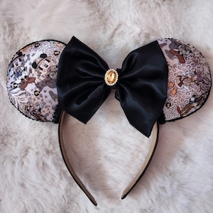 May include: Mickey Mouse ear headband with safari-themed ear designs. The ears feature a print with Mickey Mouse and friends. A large black satin bow with a gold-colored jewel is centered on the headband.