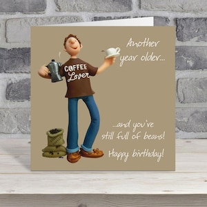 May include: A birthday card with a clay figurine of a person holding a coffee pot and a cup of coffee. The person wears a brown shirt with "COFFEE Lover" printed on it and blue jeans. The card has text that reads "Another year older... and you're still full of beans! Happy birthday!"