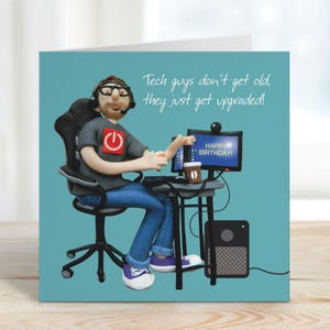 May include: Birthday card with a figurine of a person at a computer. The figurine wears glasses, a headset, and a grey t-shirt with a red power button graphic. The card reads, "Tech guys don't get old, they just get upgraded!" and "Happy Birthday!"