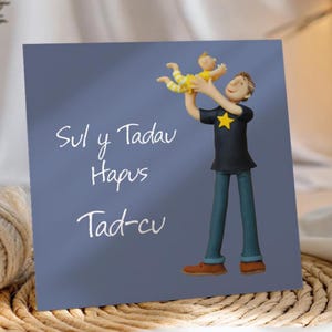 May include: A greeting card with a blue background features a cartoon illustration of a father holding a baby. The father wears a black shirt with a yellow star and blue trousers. The baby is in a yellow and white striped outfit. The card has white text.