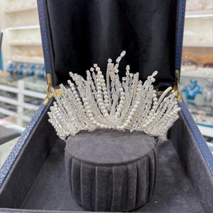 May include: A silver-toned tiara with clear crystals and faux pearls. The crown is displayed inside a blue jewelry box on a gray velvet stand. The tiara features a delicate, ornate design, perfect for weddings or special occasions.