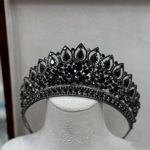 Op de afbeelding: Een zwarte tiara met een gedetailleerd ontwerp van druppel- en stervormige versieringen. De kroon wordt getoond op een witte mannequinhoofd in een bruine doos.