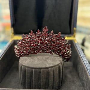 May include: A sparkling tiara with a dark red gemstone design. The crown is displayed inside a blue box with a black interior, resting on a gray velvet pedestal. The gemstones are set in a silver-toned metal.