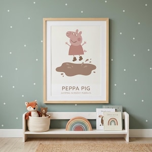 Puede incluir: Un estampado enmarcado con Peppa Pig saltando en un charco de barro. La imagen tiene un marco de madera clara y está colocada contra una pared verde pálido con lunares blancos. Una estantería blanca debajo contiene juguetes y libros.