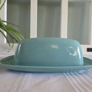 Puede incluir: Una mantequera de cerámica azul claro con tapa, sobre una base rectangular a juego. El plato está sobre un mantel blanco, con una ventana blanca y una planta verde en el fondo.