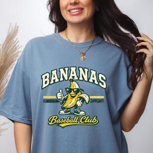 May include: Blue t-shirt with a graphic of a banana wearing sunglasses and giving a thumbs up. The text "BANANAS Baseball Club" is printed above and below the banana illustration.