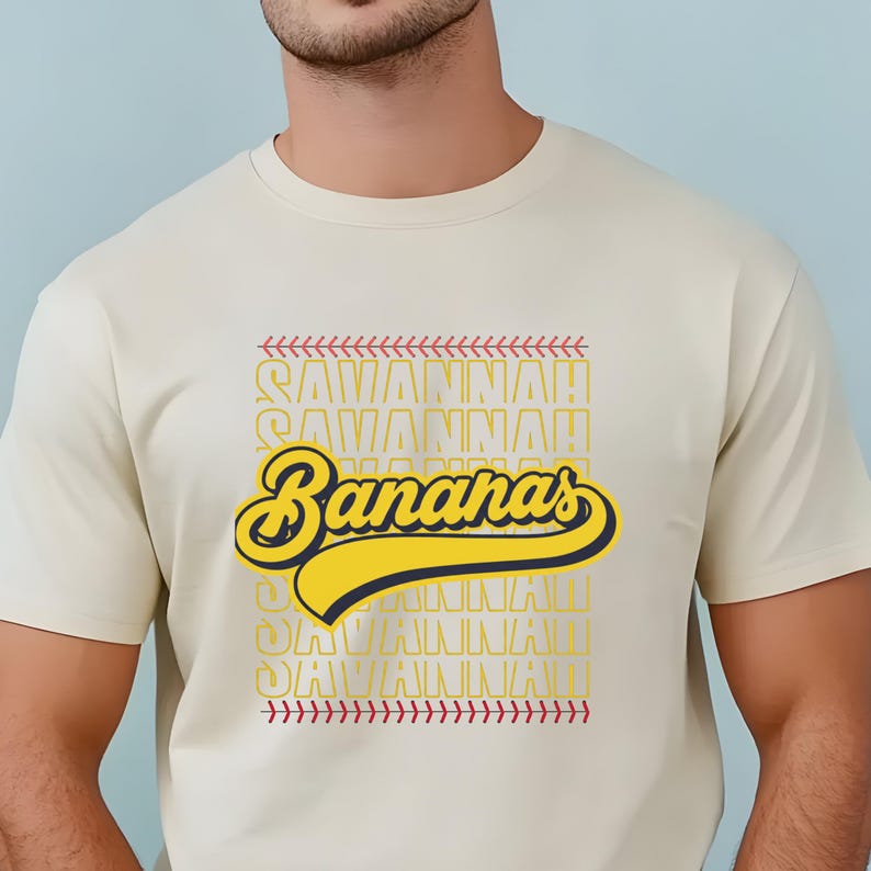 May include: Cream-colored t-shirt featuring the text "Savannah Bananas" in yellow and navy blue, with baseball-style stitching accents. The shirt's design includes the word "Savannah!" repeated in a stacked pattern.