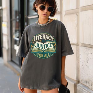 May include: A charcoal gray short-sleeve t-shirt featuring a circular graphic on the chest. The design includes an open book illustration with the text LITERACY & JUSTICE FOR ALL in cream, teal, and gold colors, set against a dark background.
