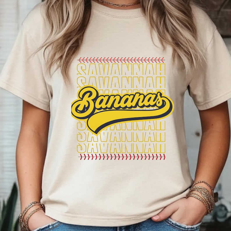 May include: Beige t-shirt with the text "Savannah Bananas" in yellow and navy blue, with baseball-style stitching and text accents. The shirt is a casual, crew neck style.