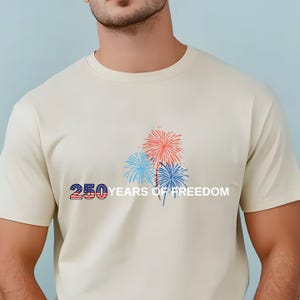 May include: A cream-colored short-sleeve t-shirt featuring a graphic of red and blue fireworks. The text 250 is displayed with a stars and stripes pattern, followed by the white text YEARS OF FREEDOM printed to the right of the fireworks.