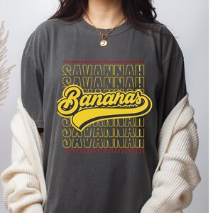 May include: Dark gray t-shirt featuring the text "Savannah Bananas" in yellow, with red accents. The design has a baseball theme. The shirt is a casual wear item.