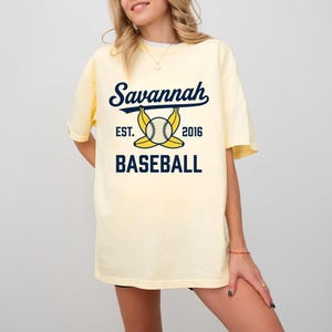 May include: A pale yellow short-sleeve t-shirt featuring a dark blue graphic print that reads Savannah BASEBALL EST. 2016, with an illustration of a baseball centered between two yellow bananas positioned horizontally across the chest area.