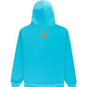 May include: A turquoise hoodie with a hood and long sleeves. An orange star-shaped logo is on the upper back. Small white dots are on the sleeves. The hoodie is made of a soft material.