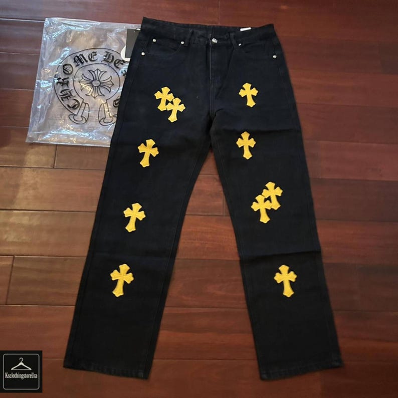 May include: Black denim jeans with multiple yellow cross appliques. The jeans have a classic five-pocket design and a straight leg fit. A clear plastic bag with a logo is visible in the background.