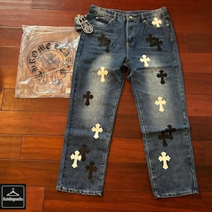 Op de afbeelding: Blauwe denim jeans met een vervaagde wassing, versierd met zwarte en witte kruisapplicaties. Een doorzichtige plastic zak met de tekst "CHROME HEARTS" is zichtbaar, samen met een klein label. De jeans hebben een klassiek vijf-zakken ontwerp.