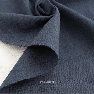May include: Close-up of a folded piece of navy blue fabric. The fabric has a textured appearance and a raw edge. The word "FABLEISM" is printed in white on the lower right corner. The fabric is likely linen or a similar material.