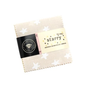 May include: A stack of beige fabric squares with white star patterns. A black band with a gold logo and text wraps around the fabric. A cream-colored label reads "starry" with text "ALEXIA MARCELLE ABEGG".