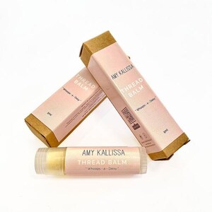 May include: Two rectangular cardboard boxes and one cylindrical tube of Amy Kallissa Thread Balm in Whoops-a-Daisy scent. The packaging is light pink with brown edges, featuring the brand name, product name, and a 5ml volume label on each item.