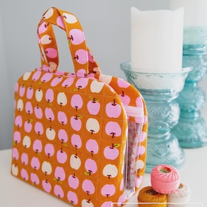 May include: A quilted orange fabric bag with a handle, featuring a repeating pattern of pink and white apples. The bag has a clear, zippered pocket on the side. Spools of thread and sewing clips are in the foreground.