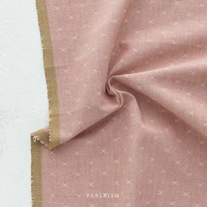 Puede incluir: Una tela de color rosa polvoriento con un patrón repetitivo de pequeñas cruces blancas bordadas. El material tiene una apariencia texturizada con un borde deshilachado y un borde de orillo color canela sólido. La palabra FABLEISM está impresa.