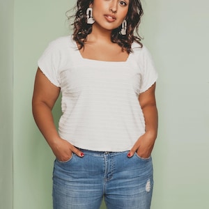 Op de afbeelding: Een vrouw draagt een witte, getextureerde top met een vierkante halslijn en een blauwe jeans met gescheurde knieën. Ze draagt witte oorbellen en heeft haar handen in haar zakken. De achtergrond is lichtgroen.