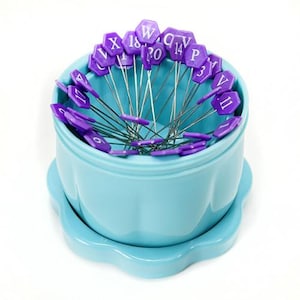 May include: A light blue ceramic container with a scalloped base holds a collection of thin metal pins. Each pin features a purple hexagonal head marked with white letters or numbers, including W, X, 18, 20, 14, P, V, 11, 17, 9, and 3.