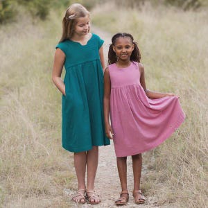 Puede incluir: Dos chicas jóvenes con vestidos. Una lleva un vestido verde azulado con mangas cortas y bolsillos, la otra un vestido rosa sin mangas. Ambos vestidos tienen la cintura fruncida y llegan a las rodillas. Están de pie en un camino de tierra.