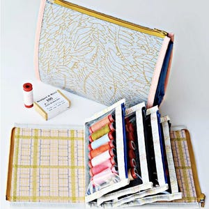 May include: A light blue sewing kit with a gold leaf pattern, pink trim, and a gold zipper. The kit is open, revealing spools of thread in various colors, a box of pins, and a spool of white thread. A second open sewing kit with a plaid pattern is also visible.