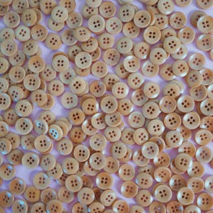 May include: A collection of light brown wooden buttons with four holes each. The buttons are round and have a slightly textured surface. They are scattered across a light pink surface, creating a visually appealing pattern. Ideal for sewing and craft projects.