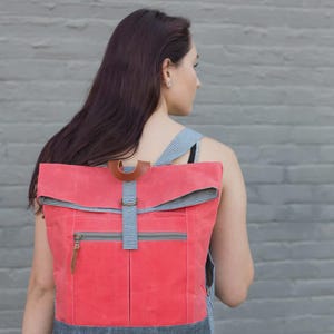 May include: A coral-colored backpack with a roll-top design. The bag features a front zippered pocket, a brown leather handle, and a blue and white checkered pattern on the flap and straps. The bottom of the bag is a dark gray denim.