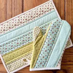 May include: A quilted oven mitt and matching hot pad set. The set features a variety of fabrics in pastel colors, including floral prints, gingham, and solid green. The edges are finished with a white binding.