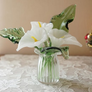 May include: A glass vase holding white calla lilies and green leaves. The clear vase has vertical ridges, and the flowers have yellow centres. The arrangement is on a table with a white lace tablecloth, with a small Christmas tree in the background.