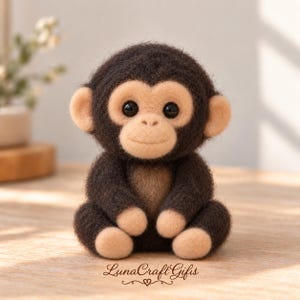 Puede incluir: Una figura de mono de lana fieltrada con pelaje marrón oscuro y cara, orejas, manos y pies de color tostado claro. El mono tiene grandes ojos negros y una nariz pequeña. El texto "LunaCraft Gifts" con un pequeño corazón está impreso debajo.