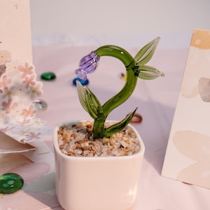 May include: A handcrafted glass flower sculpture in a white ceramic pot. The flower has a green stem and leaves, with a purple bud. The pot is filled with small pebbles. Decorative glass beads are scattered around the pot.