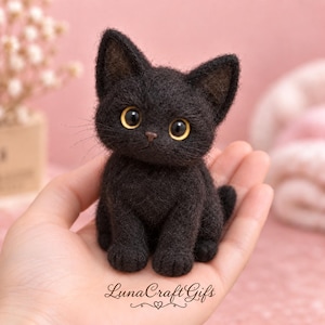 Peut inclure: Une petite figurine de chat noir fabriquée à la main en laine feutrée. Le chat a de grands yeux jaunes brillants, des oreilles pointues et un aspect texturé et duveteux. Le texte "LunaCraftGifts" est imprimé en cursive au bas de l'image.